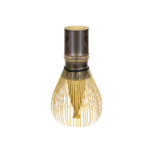 A traditional Japanese bamboo whisk (chashaku) with a dark brown bamboo top and fine gold-colored whisking bristles, isolated on a white background.